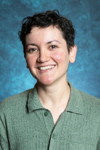 Picture of Dr. Rio Lopez Director of Latin American Programs. The image is a headshot of a person smiling against a blue mottled backdrop. The person has short, curly dark hair and is wearing a light green, ribbed, knit collared shirt with a button-down front. There are three visible buttons on the shirt. The backdrop is a textured mix of darker and lighter shades of blue, adding depth to the background without distracting from the subject.